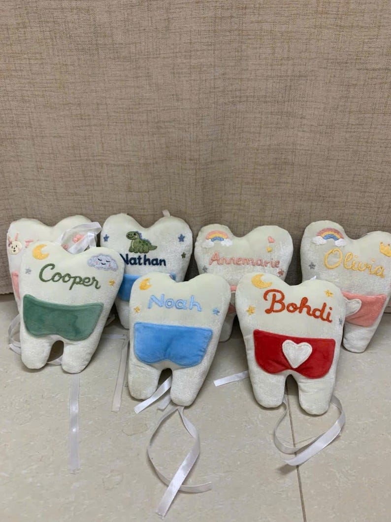 May include: A collection of plush tooth-shaped pillows in off-white fabric, each featuring embroidered names including Cooper, Nathan, Noah, Annemarie, Bohdi, and Olivia. The pillows have small colourful pockets and attached white ribbons.