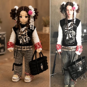 May include: A crocheted doll with dark hair styled in pigtails, wearing a black vest over a white long-sleeved shirt, gray pants, and sneakers. The doll carries a black tote bag and is accessorized with a belt and wrist cuffs. The doll's outfit mirrors the style of the person standing next to it.