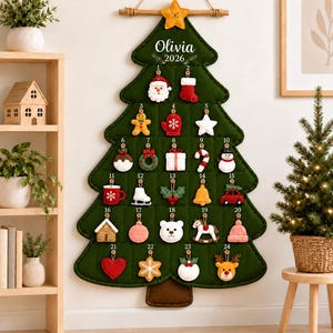 Peut inclure: Un calendrier de l'Avent mural en feutrine vert foncé en forme de sapin de Noël avec une tige en bois et une cordelette. Il porte le nom Olivia et l'année 2026, avec 24 boutons numérotés supportant des ornements en feutrine comme un Père Noël.