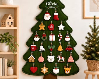 May include: A dark green felt Christmas tree wall hanging advent calendar with a wooden dowel and hanging cord. It features the name Olivia and the year 2026, with 24 numbered buttons holding individual felt ornaments like Santa, a stocking, and a reindeer.