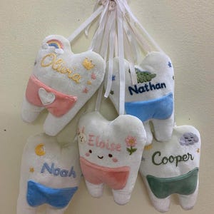 May include: Five plush white tooth-shaped pillows hanging from white ribbons. Each pillow features embroidered names: Olivia, Nathan, Eloise, Noah, and Cooper. They include colourful accents like a rainbow, crown, stars, flowers, and clouds, with small fabric pockets in pink, blue, or green.
