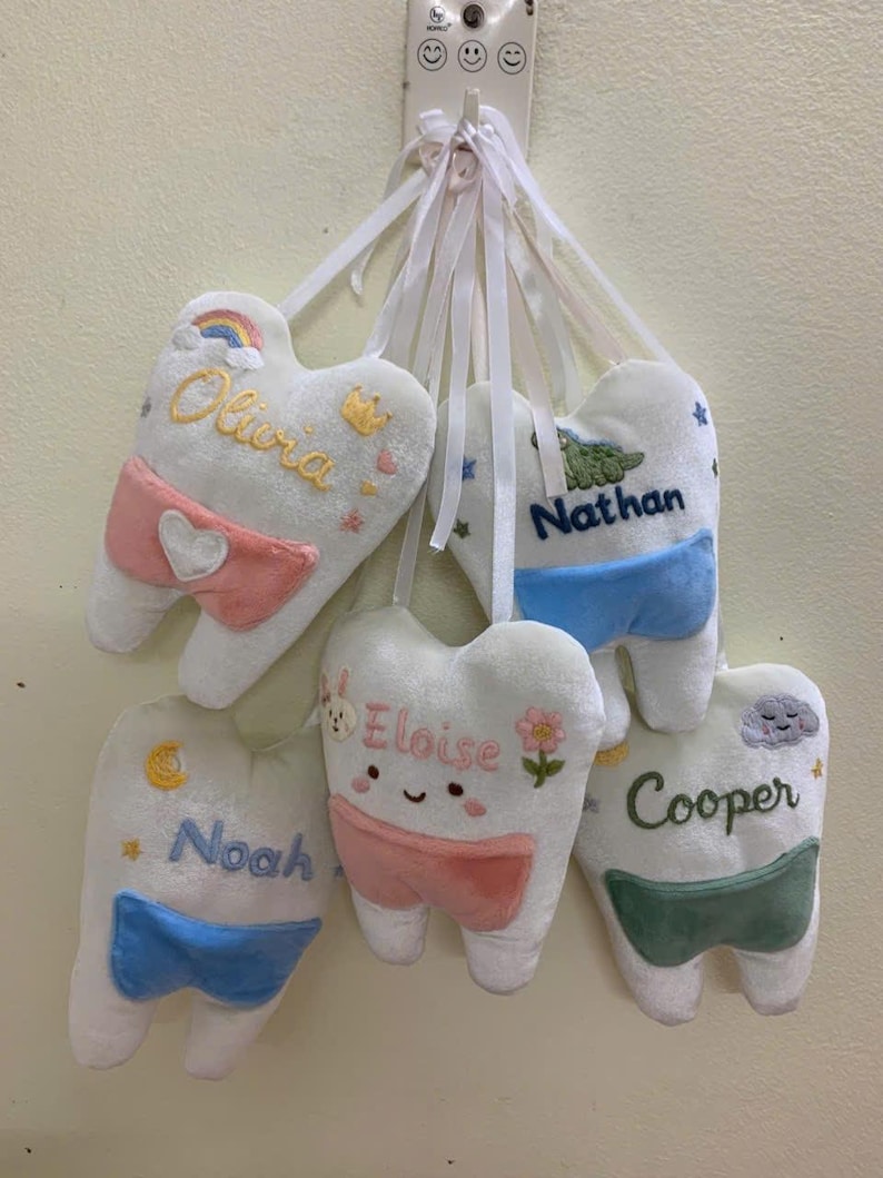 May include: Five plush white tooth-shaped pillows hanging from white ribbons. Each pillow features embroidered names: Olivia, Nathan, Eloise, Noah, and Cooper. They include colourful accents like a rainbow, crown, stars, flowers, and clouds, with small fabric pockets in pink, blue, or green.