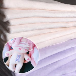 K&ouml;nnte beinhalten: Ein Stapel weicher, hellfarbener Stoffe mit einer Pl&uuml;schstruktur. Ein kreisf&ouml;rmiger Einsatz zeigt ein Pl&uuml;schkaninchen mit rosa Akzenten, einschlie&szlig;lich Ohren, Bandagen und herzf&ouml;rmigen Pfoten. Das Kaninchen hat ein blaues Korsettmuster auf der Brust.