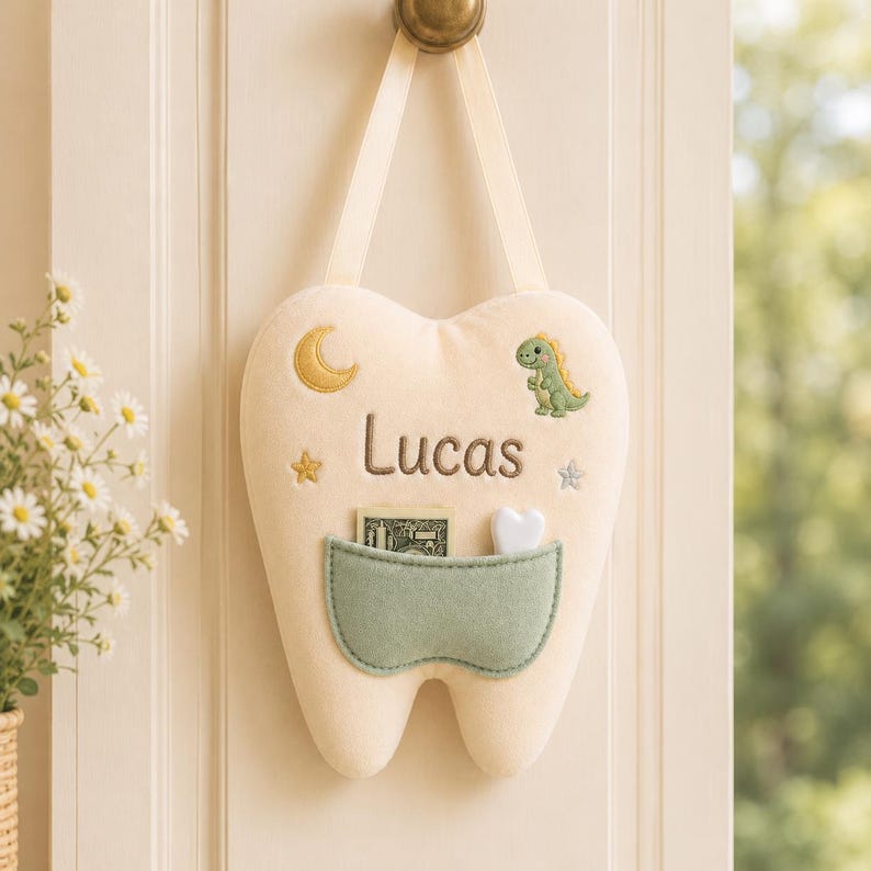May include: A cream-coloured plush tooth-shaped wall hanging with a ribbon loop for suspension. It features an embroidered yellow crescent moon, a green dinosaur, stars, and the name Lucas in brown text, with a sage green pocket holding money.
