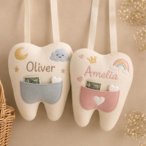 May include: Two cream-coloured felt tooth-shaped pillows with hanging straps. One features the name Oliver, a yellow moon, and a blue cloud above a blue pocket. The other features the name Amelia, a yellow crown, and a rainbow above a pink pocket.