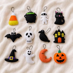 May include: A set of twelve felt Halloween ornaments featuring a candy corn, a black cauldron with green bubbles, a white mummy, a black cat, a bat, a skull, a black crow, a haunted house, a witch hat, a white ghost, an orange moon, and a pumpkin.