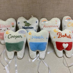 May include: A collection of seven plush tooth-shaped pillows in white fabric. Each pillow features embroidered names including Eloise, Nathan, Annemarie, Olivia, Cooper, Noah, and Bohdi, along with colourful embroidered icons and attached fabric pockets.