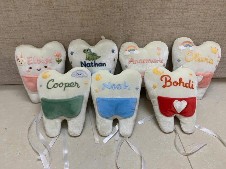 May include: A collection of seven plush tooth-shaped pillows in white fabric. Each pillow features embroidered names including Eloise, Nathan, Annemarie, Olivia, Cooper, Noah, and Bohdi, along with colourful embroidered icons and attached fabric pockets.