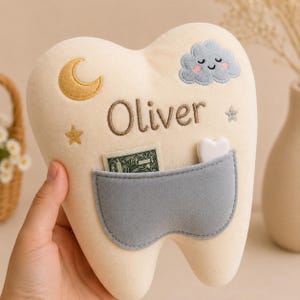 May include: A cream-coloured plush tooth pillow featuring the embroidered name Oliver, a gold crescent moon, a grey smiling cloud, and two small stars. A grey pocket on the front holds a small white heart-shaped object and a piece of paper currency.