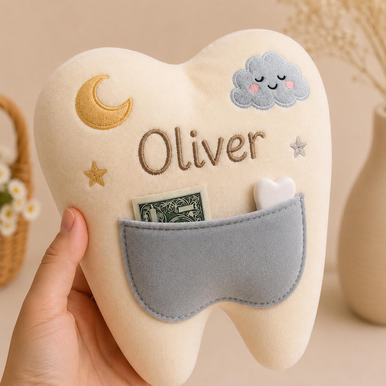 May include: A cream-coloured plush tooth pillow featuring the embroidered name Oliver, a gold crescent moon, a grey smiling cloud, and two small stars. A grey pocket on the front holds a small white heart-shaped object and a piece of paper currency.