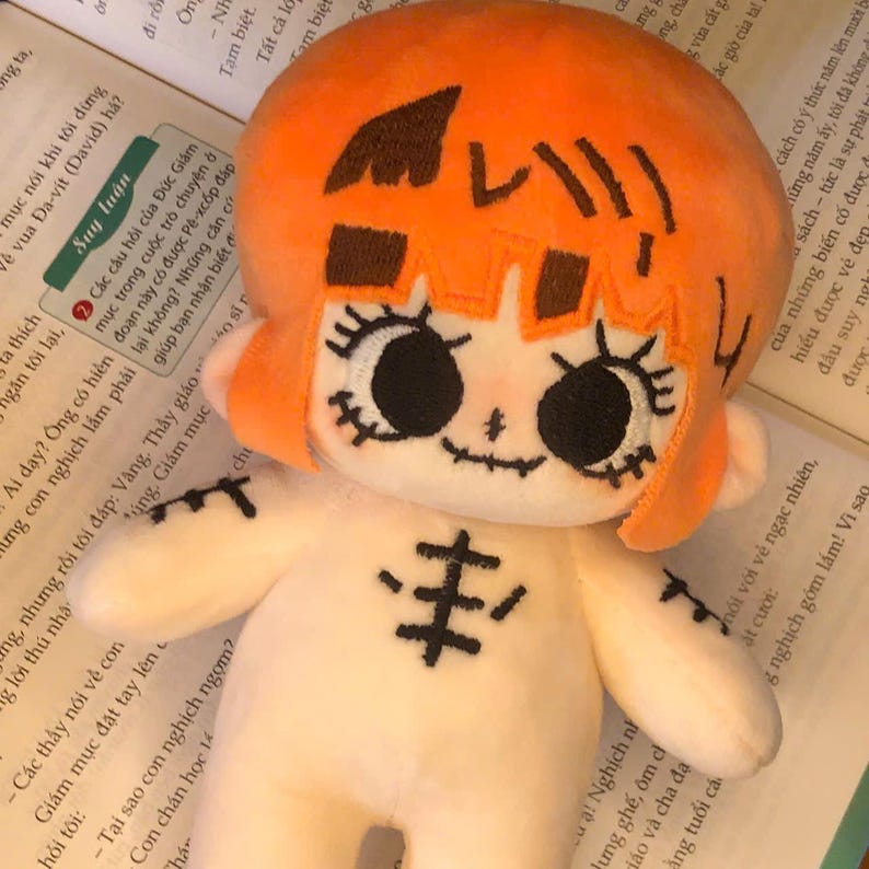 K&ouml;nnte beinhalten: Pl&uuml;schpuppe mit orangefarbenem Haar und aufgestickten Gesichtsz&uuml;gen, einschlie&szlig;lich gro&szlig;er Augen und eines aufgestickten L&auml;chelns. Die Puppe hat einen wei&szlig;en K&ouml;rper mit einem aufgestickten Design auf der Brust und liegt auf einem Buch.