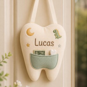 May include: A cream-coloured plush tooth-shaped pillow with a hanging ribbon. It features an embroidered crescent moon, a green dinosaur, two stars, and the name Lucas in brown thread. A sage green pocket holds a banknote and a small white heart.