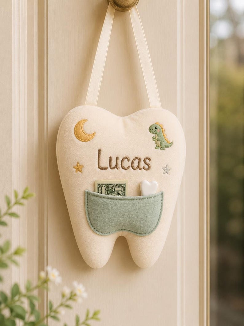 May include: A cream-coloured plush tooth-shaped pillow with a hanging ribbon. It features an embroidered crescent moon, a green dinosaur, two stars, and the name Lucas in brown thread. A sage green pocket holds a banknote and a small white heart.