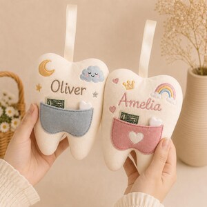 May include: Two plush tooth-shaped pillows with hanging ribbons. One is embroidered with the name Oliver, a moon, stars, and a blue cloud, featuring a blue pocket. The other is embroidered with Amelia, a crown, hearts, and a rainbow, with a pink pocket.