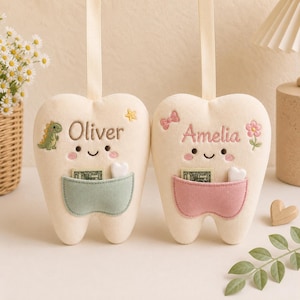 May include: Two cream-coloured plush tooth-shaped pillows with hanging ribbons. One features the name Oliver, a green dinosaur, and a sage green pocket. The other features the name Amelia, a pink bow, a flower, and a dusty pink pocket.