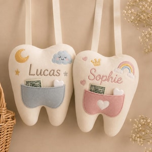 May include: Two cream-coloured tooth-shaped plush pillows with hanging ribbons. The left pillow features a blue pocket, a yellow moon, and the name Lucas. The right pillow features a pink pocket, a gold crown, a rainbow, and the name Sophie.