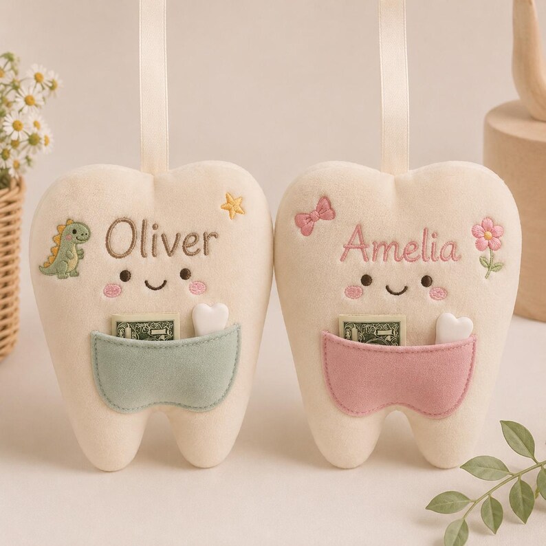 May include: Two cream-coloured plush tooth-shaped pillows with hanging ribbons. One features the name Oliver, a green dinosaur, and a sage green pocket. The other features the name Amelia, a pink bow, a flower, and a dusty pink pocket.