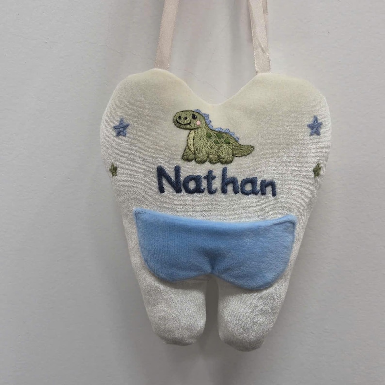 May include: A white tooth-shaped plush hanging pouch with a light blue pocket. The front features an embroidered green dinosaur, small blue and green stars, and the name Nathan stitched in dark blue thread. It includes a white fabric hanging loop.