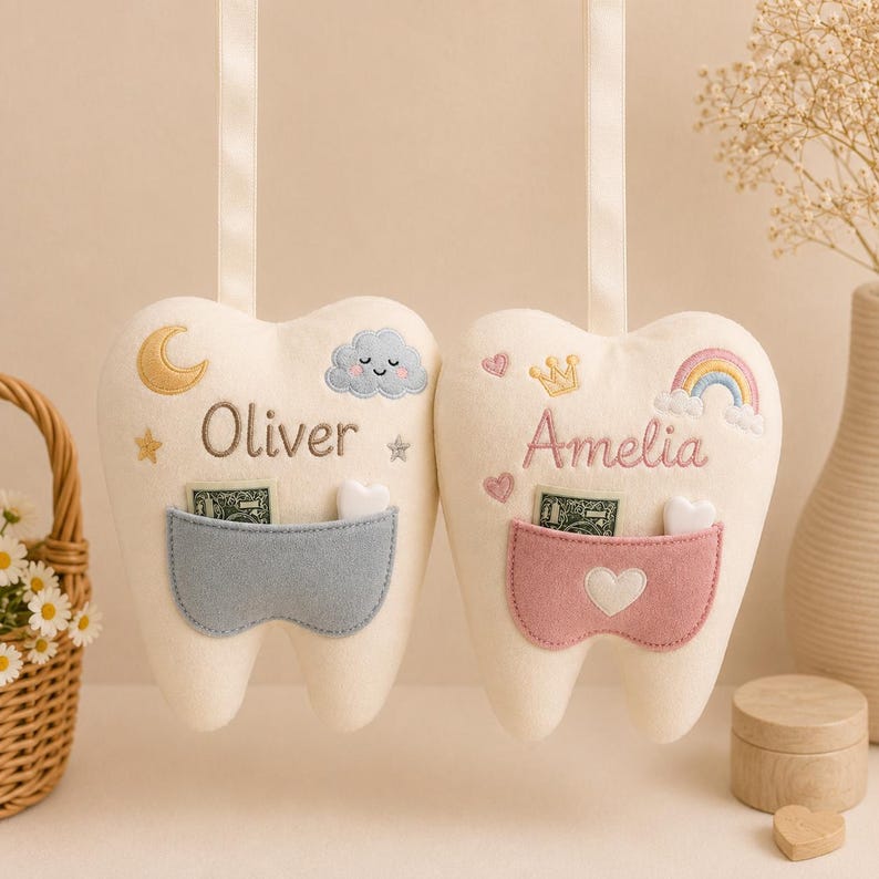 May include: Two cream-coloured felt tooth-shaped cushions with hanging ribbons. One features a blue pocket, a moon, a cloud, and the name Oliver. The other has a pink pocket, a heart, a crown, a rainbow, and the name Amelia. Both contain small notes.