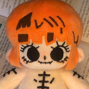 K&ouml;nnte beinhalten: Eine Pl&uuml;schpuppe mit einem orangefarbenen, k&uuml;rbisf&ouml;rmigen Kopf und einem wei&szlig;en K&ouml;rper. Die Puppe hat gestickte schwarze Augen, einen gen&auml;hten Mund und ein Skelettmuster auf dem K&ouml;rper. Das Haar der Puppe ist orange und das Gesicht ist wei&szlig;.