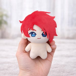 K&ouml;nnte beinhalten: Eine Pl&uuml;schpuppe mit roten Haaren und blauen Augen wird in einer Hand gehalten. Die Puppe hat einen cremefarbenen K&ouml;rper und ein einfaches, l&auml;chelndes Gesicht. Das Haar der Puppe ist stachelig frisiert.