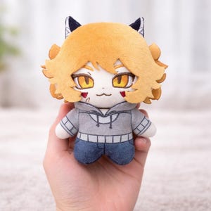 K&ouml;nnte beinhalten: Eine Pl&uuml;schpuppe mit katzenartigem Aussehen, mit orangefarbenem Haar, gelben Augen und einem grauen Kapuzenpullover mit blauen Jeans. Die Puppe hat kleine schwarze Ohren und rote Markierungen auf den Wangen. Sie wird in einer Hand gehalten.