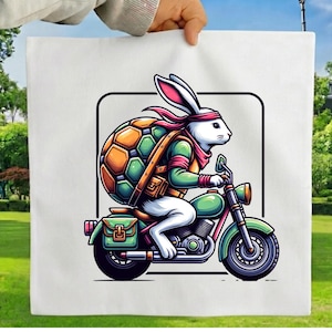 May include: A white tote bag featuring a graphic illustration of a white rabbit wearing a red headband and a green and orange turtle shell backpack while riding a green motorcycle with a side saddlebag, all enclosed in a black square frame.