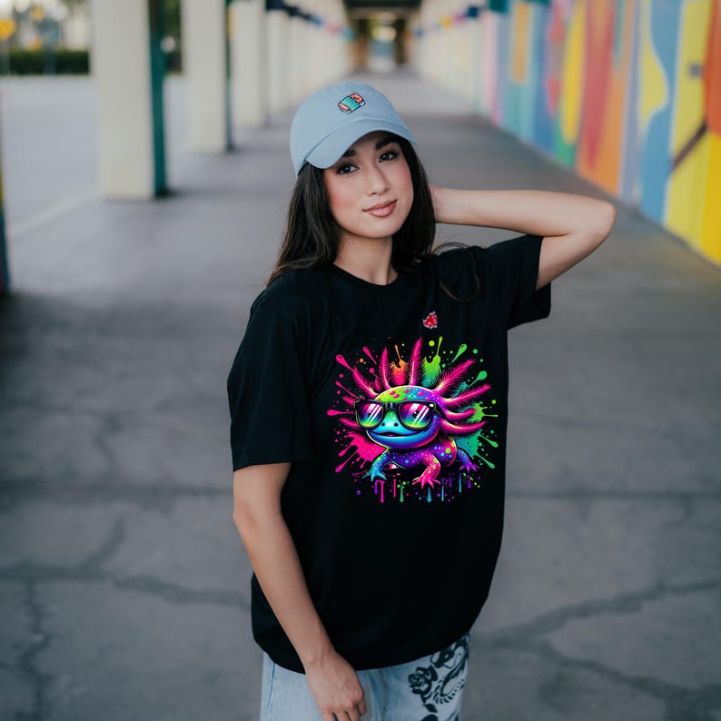 Pu&ograve; includere: T-shirt nero con un colorato axolotl con occhiali da sole, su uno sfondo di vivaci schizzi di vernice. Il design dell'axolotl include sfumature di blu, verde, rosa e viola. &Egrave; visibile anche un berretto da baseball azzurro.