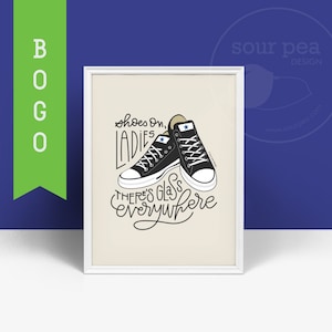 May include: A white framed print with a beige background featuring a black and white illustration of two black sneakers with the text "shoes on, ladies there's glass everywhere".