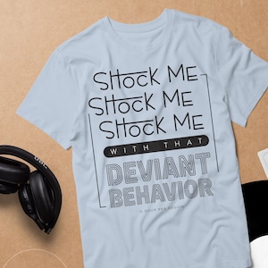 May include: Light blue t-shirt with the text "Shock Me" repeated three times, and "With That Deviant Behavior" in a bold, outlined font. The shirt is displayed with headphones, a cassette tape, and a vinyl record.