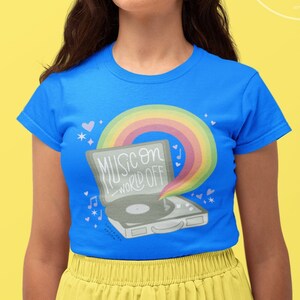 May include: A woman wearing a blue t-shirt with a rainbow and a record player graphic. The graphic says "Music on World off". She is wearing a yellow skirt.