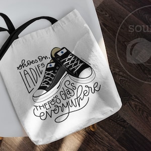 May include: White canvas tote bag with black straps. The bag features a black and white illustration of two sneakers with the text "shoes on, ladies there's glass everywhere".