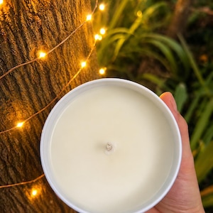 May include: A round, white metal container filled with smooth, off-white wax featuring a single centred cotton wick. The container is held in a palm, showcasing the flat, circular surface of the candle wax from a top-down perspective.