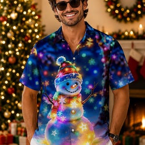 May include: A short-sleeved button-down shirt featuring a vibrant, colorful graphic print of a snowman wearing a striped hat and scarf. The design includes glowing star patterns and bokeh light effects against a deep blue background.