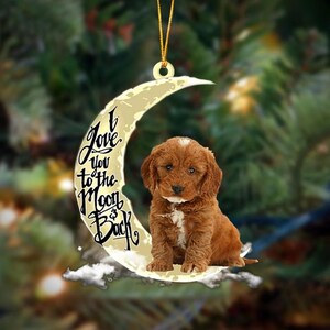 May include: A crescent moon-shaped ornament featuring a brown puppy with a white chest patch. The moon is cream-coloured with black script text that reads I Love you to the Moon & Back. A gold string is attached to the top for hanging purposes.