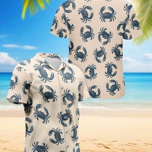 Puede incluir: Dos camisas de manga corta con botones y fondo color crema. Las camisas están cubiertas con un patrón repetido de ilustraciones de cangrejos azul oscuro. Las camisas se exhiben en una playa con cielo azul y palmeras al fondo.