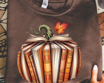 May include: Brown sweatshirt featuring a pumpkin design made of books. The pumpkin has a green stem and a red and orange leaf. The books are various shades of brown and orange.