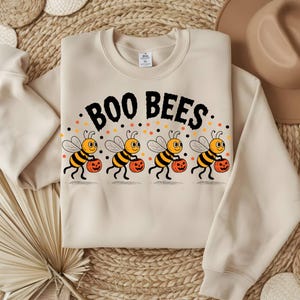 May include: A cream-coloured sweatshirt featuring the words "BOO BEES" in black. Below the text are four cartoon bees, each carrying an orange pumpkin. The bees have yellow and black striped bodies, wings, and antennae. Small orange and black dots are above the bees.