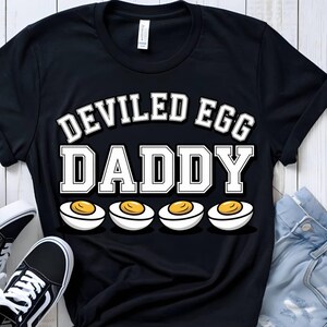 Könnte beinhalten: Schwarzes T-Shirt mit dem Text "DEVILED EGG DADDY" in weißen Blockbuchstaben. Darunter befinden sich vier Cartoon-Eier mit gelben Eigelben. Das T-Shirt hat einen lässigen Stil.