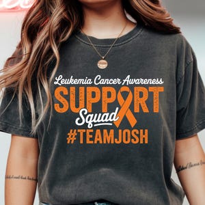 May include: Dark gray t-shirt with the text "Leukemia Cancer Awareness SUPPORT Squad #TEAMJOSH" in white and orange. The shirt features an orange awareness ribbon.