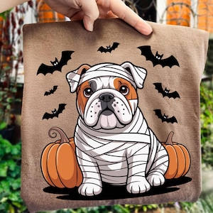 May include: A brown Halloween-themed pillow featuring a bulldog dressed as a mummy, with orange pumpkins and black bat silhouettes. The bulldog has brown and white fur and is wrapped in white bandages.