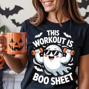 May include: Black t-shirt with a cartoon ghost lifting weights, wearing sunglasses, and the text "THIS WORKOUT IS BOO SHEET." An orange mug with a jack-o'-lantern face is held in the hand.