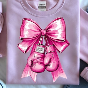 May include: A light purple sweatshirt featuring a large pink bow and two pink boxing gloves. The gloves have the words "BOXING HOPE" printed on them. The sweatshirt is made of a soft material.