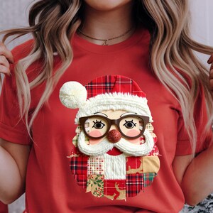 Puede incluir: Camiseta roja con un diseño de cara de Papá Noel. La cara de Papá Noel incluye un gorro a cuadros rojos y blancos, gafas marrones, barba blanca y nariz roja con purpurina. La cara de Papá Noel está hecha de varios parches de tela.