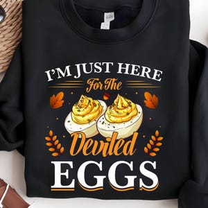 Op de afbeelding: Zwarte sweatshirt met de tekst "I'M JUST HERE For The Deviled EGGS" in wit en oranje. De afbeelding toont twee gevulde eieren met gele vulling en oranje en bruine herfstbladeren.
