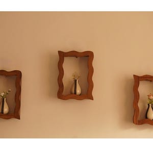 May include: Three wooden wall shelves with a wavy frame design. Each shelf holds a small white vase with a black stripe and a few flowers. The shelves are mounted on a beige wall, creating a decorative display.