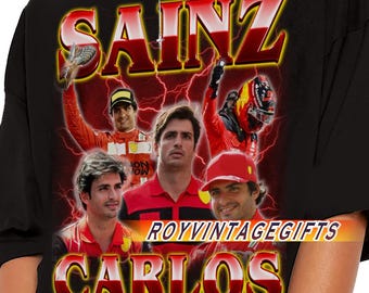 Op de afbeelding: Zwart T-shirt met een afbeelding van Carlos Sainz, met de tekst "SAINZ" en "CARLOS" in een rood en geel ontwerp. Het shirt bevat afbeeldingen van Carlos Sainz in een race-uniform. De tekst "ROYVINTAGEGIFTS" is ook zichtbaar.