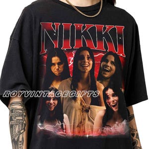 May include: A black short-sleeve t-shirt featuring a graphic print with the name NIKKI in large red letters at the top. The design includes five different photographic portraits of a woman with long dark hair, set against a red background.