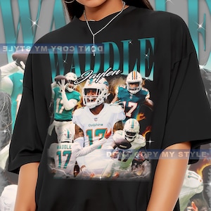 Puede incluir: Camiseta negra con un gráfico del jugador de fútbol americano Jaylen Waddle con el uniforme de los Miami Dolphins. El diseño incluye el nombre "WADDLE" en color teal y el número 17. La camiseta tiene un aspecto vintage.