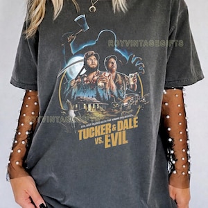 May include: Dark gray t-shirt featuring a movie poster design for "Tucker & Dale vs. Evil." The graphic includes two men, a house, and the film's title in a distressed font. The text "EVIL JUST MESSED WITH THE WRONG HILLBILLIES" is also present.